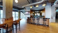This single-floor clubhouse delivers a distinctive industrial-chic aesthetic, blending raw architectural elements with warm, inviting finishes throughout its open layout. Exposed concrete columns, visible ductwork, and an open ceiling structure define the space's character, while light-toned hardwood flooring and rich wood paneling provide warmth and contrast.

The social hub of the clubhouse centers around a built-in bar framed in natural wood paneling, complete with a dark countertop, bar seating with chrome stools, and overhead pendant lighting. A wall-mounted television nearby adds a casual entertainment element to the bar area. High-top communal tables and bar-height chairs offer additional seating for gathering and conversation.

Adjacent to the bar, an open lounge area features leather and upholstered seating arranged around a bold graphic rug with a starburst pattern in navy and yellow. This seating zone is bathed in abundant natural light, courtesy of large floor-to-ceiling windows and glass doors that line the perimeter walls and open to an outdoor space. Vibrant yellow accent walls punctuate the concrete columns, adding energy and personality to the room.

The combination of industrial structure, warm wood details, lively color accents, and generous natural light creates a versatile, welcoming environment well-suited for socializing, relaxing, and community gatherings.