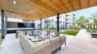 Located in Annapolis, MD, this thoughtfully designed second courtyard offers a refined outdoor gathering space set within a contemporary multi-story building complex. The courtyard is anchored by a striking covered pavilion featuring a warm slatted wood ceiling, overhead infrared heaters, and bold black steel columns — providing shade and comfort throughout the seasons. Beneath the pavilion, plush wicker lounge seating with colorful accent pillows surrounds a central coffee table, while a dedicated dining area and an outdoor television make it ideal for both relaxed lounging and social gatherings. An outdoor kitchen or bar station is integrated into the pavilion's layout, adding functional amenity to the space. Beyond the pavilion, a broad concrete pathway stretches the length of the courtyard, flanked by lush manicured lawn panels and richly planted garden beds featuring ornamental grasses, flowering perennials, and young trees that lend vertical interest. Intimate seating vignettes with modern bistro tables and chairs are positioned throughout, offering quiet retreats along the walkway. The surrounding multi-story buildings, with their balconies and large-paned windows, frame the courtyard and contribute to its sense of enclosure and community. The overall ambiance is fresh, modern, and inviting — a well-appointed outdoor environment designed for everyday enjoyment.