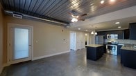 This single-floor space showcases a compelling blend of industrial and modern design elements throughout its open-concept layout. The corrugated metal ceiling serves as a defining architectural feature, complemented by an exposed silver HVAC duct that reinforces the industrial aesthetic. A ceiling fan with integrated lighting is centrally positioned, while recessed lighting provides additional illumination throughout.

The polished concrete floors extend seamlessly across the entire space, offering a sleek, low-maintenance surface that ties the industrial theme together cohesively. The living area is generously proportioned with neutral beige walls, providing a clean backdrop and ample flexibility for furniture arrangement.

The kitchen occupies the right side of the space and features dark cabinetry paired with a light-colored countertop island, creating a striking contrast. Stainless steel appliances add a contemporary touch, while pendant lighting hangs above the island, adding warmth and definition to the cooking area.

White interior doors lead to additional rooms beyond the main living space. The overall ambiance is modern and urban, balancing raw industrial materials with functional, well-designed living spaces.