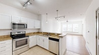 This single-floor unit is designed with a clean, contemporary aesthetic and a bright, open layout. The kitchen serves as a functional centerpiece, featuring white shaker-style cabinetry, granite countertops with a warm golden tone, and a full suite of stainless steel appliances including a refrigerator, range, over-the-range microwave, and dishwasher. A peninsula with an extended countertop provides additional workspace and separates the kitchen from the living area. The kitchen is finished with neutral tile flooring set in a diagonal pattern and illuminated by a combination of pendant lights and track lighting.

The living area extends naturally from the kitchen and is bathed in natural light from large windows and a glass-paneled exterior door. Light hardwood flooring adds warmth to this open space. Bedrooms are carpeted and feature windows with horizontal blinds, allowing for adjustable natural light and views of the surrounding area. One bedroom includes a wide three-panel window arrangement that enhances the sense of openness.

The bathroom is finished with granite-topped white vanity cabinetry, a tiled tub-shower combination, and coordinating tile flooring, maintaining the consistent design language found throughout the unit. An entry foyer with tile flooring connects the main door to the rest of the space.