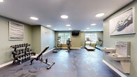 Located in Auburn, Maine, this fitness center offers a comprehensive and well-appointed exercise facility across a single floor. The exercise room is the heart of the space, finished with a durable, marbled epoxy-style floor and sage green walls that create a clean, energizing atmosphere. Recessed circular lighting throughout the ceiling provides bright, even illumination across the entire area. Large windows along the walls invite natural light and offer views of the surrounding greenery, enhancing the open feel of the room. The exercise room is equipped with multiple treadmills, an elliptical machine, a stationary bike, an adjustable weight bench, and a tiered dumbbell rack — providing a well-rounded selection of both cardio and strength training equipment. A wall-mounted flat-screen television adds convenience for entertainment during workouts. The entry area includes a dual-basin water fountain and wall art, adding a welcoming touch to the space. A dedicated bathroom adjoins the main exercise area, featuring a classic black-and-white dotted tile floor, pedestal sink with mirror, ADA-compliant grab bars, and clean sage green walls consistent with the overall design aesthetic. The facility also includes an accessible exit door, ensuring both safety and ease of access throughout.