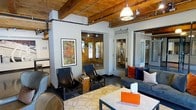 This single-floor space masterfully combines industrial character with modern comfort. Overhead, rich exposed wood beams and joists span the ceiling alongside visible black iron piping, lending a raw, warehouse-inspired aesthetic that is balanced by crisp white walls and carefully curated furnishings. Pendant lighting fixtures hang from the ceiling, casting a warm, inviting glow throughout the open interior.

The main lounge area is thoughtfully arranged with a mix of seating options, including a plush gray sofa accented with bold orange and navy throw pillows, sleek black leather sling chairs, and a nailhead-trimmed accent chair. A patterned area rug anchors the seating arrangement, adding warmth to the polished concrete floors beneath. A wooden side table and a central coffee table complete the setup, offering both style and functionality.

Colorful framed artwork decorates the walls, adding a creative, vibrant touch to the space. Glass-paneled doors and partitions allow natural light to flow freely, contributing to an open, airy feel. The layout suggests a professional yet welcoming environment, well-suited for a creative office, coworking space, or client-facing business. The overall ambiance is sophisticated, warm, and energetic.