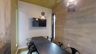 This thoughtfully designed single-floor space serves as a functional and visually striking meeting or conference room. The interior showcases a compelling blend of modern and contemporary design, anchored by a striking accent wall clad in warm, natural wood planking that adds texture and depth to the room. Contrasting crisp white walls provide a clean backdrop, while a bold gold-painted ceiling introduces an unexpected pop of color and elegance overhead. Artistic wire-frame pendant lights hang from the ceiling, serving as sculptural focal points that cast a warm, ambient glow throughout the space. A wall-mounted flat-screen display is centrally positioned, making it ideal for presentations and collaborative discussions. The room is furnished with a dark wood conference table surrounded by sleek black chairs, creating a cohesive and professional aesthetic. A narrow vertical window along the wood-accented wall allows natural light to filter in, enhancing the overall ambiance. The combination of natural materials, curated lighting, and modern furnishings gives this space a refined yet inviting character, well-suited for productive meetings or creative gatherings.