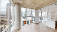 This well-appointed two-bedroom, two-bathroom residence showcases a contemporary urban design characterized by exposed concrete ceilings and expansive floor-to-ceiling windows that flood the interior with natural light. The open living area is anchored by dramatic views of the surrounding cityscape and nearby hillside, with large glazed panels extending across multiple walls to maximize visual connectivity with the outdoors.

The kitchen is a standout feature, equipped with a substantial quartz-topped island, gray shaker-style cabinetry, a subway tile backsplash, stainless steel appliances, and pendant lighting. It connects seamlessly to the living area, reinforcing the open-plan layout.

Light wood-look flooring runs throughout the unit, complementing the neutral white walls and concrete architectural elements. A clean, bright hallway leads to the two bedrooms, each featuring exposed concrete ceilings, natural light, and access to closet space. Two dedicated closets provide practical storage.

The primary bathroom offers a long vanity with gray cabinetry, a white quartz countertop, and built-in shelving, with direct access to the primary bedroom. The overall ambiance is modern, airy, and urban.