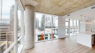 This well-appointed two-bedroom, two-bathroom residence showcases a contemporary urban design characterized by exposed concrete ceilings and expansive floor-to-ceiling windows that flood the interior with natural light. The open living area is anchored by dramatic views of the surrounding cityscape and nearby hillside, with large glazed panels extending across multiple walls to maximize visual connectivity with the outdoors.

The kitchen is a standout feature, equipped with a substantial quartz-topped island, gray shaker-style cabinetry, a subway tile backsplash, stainless steel appliances, and pendant lighting. It connects seamlessly to the living area, reinforcing the open-plan layout.

Light wood-look flooring runs throughout the unit, complementing the neutral white walls and concrete architectural elements. A clean, bright hallway leads to the two bedrooms, each featuring exposed concrete ceilings, natural light, and access to closet space. Two dedicated closets provide practical storage.

The primary bathroom offers a long vanity with gray cabinetry, a white quartz countertop, and built-in shelving, with direct access to the primary bedroom. The overall ambiance is modern, airy, and urban.