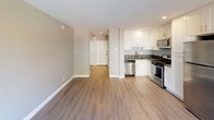 This single-floor unit offers a clean, modern aesthetic throughout, characterized by light wood-style vinyl flooring, neutral gray and white wall tones, and recessed lighting. The open-concept living area provides a generous amount of space, anchored by a large window that invites natural light and frames views of surrounding greenery. A wall-mounted air conditioning unit keeps the space comfortable year-round.

The kitchen is thoughtfully designed with crisp white shaker-style cabinetry, dark quartz countertops, and a full suite of stainless steel appliances, including a gas range, over-the-range microwave, dishwasher, and refrigerator. A stainless steel undermount sink completes the functional workspace, making it well-suited for everyday cooking.

Sliding mirrored closet doors are visible near the hallway, maximizing both storage and the sense of space. The layout flows efficiently from the main living area through a corridor that connects to the bathroom.

The bathroom is finished in a clean, minimal style featuring a full-size soaking tub with a shower and curtain rod, a toilet, and a wall-mounted cabinet with a round mirror. The gray wall tones carry throughout, providing a cohesive and modern feel across the entire unit.