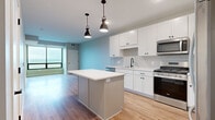 This single-floor residence is designed with a clean, contemporary aesthetic throughout. The kitchen serves as a central hub of the layout, showcasing white shaker-style cabinetry, a classic subway tile backsplash, and a full suite of stainless steel appliances including a gas range, built-in microwave, dishwasher, and French door refrigerator. A central island with a crisp white countertop provides additional workspace and storage, illuminated overhead by industrial-style pendant lights. Light hardwood flooring extends throughout the main living areas, complementing the neutral gray and soft blue wall tones.

The living area benefits from large floor-to-ceiling windows fitted with black frames, drawing in abundant natural light and offering open views of the surrounding landscape. An adjoining bedroom continues the light, airy palette with plush carpeting, a ceiling fan, and its own set of expansive windows framing scenic outdoor views.

Storage is well-considered throughout, with a walk-in closet featuring multi-tier wire shelving and a separate closet offering additional hanging and shelf space. A connected hallway with hardwood flooring links the bedroom area to the bathroom, which includes built-in cabinetry with black hardware. The overall ambiance is bright, functional, and move-in ready.