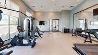 This fitness center offers a comprehensive workout environment within a thoughtfully designed single-room layout. The space is characterized by soft teal and neutral-toned walls that create a calm, focused atmosphere, complemented by light vinyl flooring that is both durable and easy to maintain. Elevated windows along multiple walls flood the room with abundant natural light, while roller shades allow for adjustable brightness. Ceiling fans provide consistent air circulation throughout the workout area.

The cardio section features multiple treadmills, an elliptical trainer, a stationary bike, and a rowing machine, providing a full range of aerobic exercise options. A dedicated strength training area includes a multi-function cable machine with adjustable bench, along with a dumbbell rack stocked with a variety of free weights. A large wall-mounted mirror expands the sense of space visually, while flat-screen televisions mounted nearby keep users entertained during workouts.

A textured accent wall provides visual interest near the room's entrance, where a wall-mounted TV and open storage unit offer added convenience. Recessed lighting and ceiling fans complete the clean, modern aesthetic. The overall design balances functionality with a polished, welcoming environment suited for a variety of fitness routines.