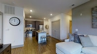 This single-floor space showcases a thoughtfully designed open-concept layout that seamlessly connects the kitchen and living area. The kitchen is a standout feature, equipped with stainless steel appliances including a range, microwave, and refrigerator, complemented by dark espresso cabinetry that provides generous storage. A light-toned countertop island anchors the kitchen, offering additional workspace and casual bar seating with two stools — ideal for dining or entertaining. The flooring throughout is a warm-toned hardwood, lending consistency and a clean aesthetic across the entire space. Recessed lighting is installed throughout the ceiling, ensuring even, well-distributed illumination. The living area is open and inviting, flowing naturally from the kitchen without visual obstruction. A set of double panel doors adds architectural interest and provides access to an adjoining space. The overall color palette is neutral and modern, with white walls, gray accents, and dark cabinetry creating a balanced contrast. The layout maximizes functionality while maintaining an airy, uncluttered feel suited to everyday living.