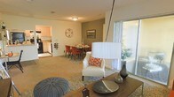 This single-story residence showcases an open-concept floor plan that seamlessly connects the living, dining, and kitchen areas into one cohesive and functional space. The living area is carpeted and comfortably proportioned, anchored by a large sliding glass door that introduces generous natural light and provides direct access to an outdoor area, offering pleasant views of surrounding greenery. An accent wall in a warm taupe-green tone adds visual depth to the dining area, which is defined by a area rug and features a mid-century style dining table surrounded by bold red upholstered chairs — a striking focal point within the space. The kitchen, visible from the main living area, is appointed with light wood cabinetry, black appliances, and a practical layout suited for everyday use. Consistent carpeting flows throughout the main living spaces, contributing to a warm and cohesive aesthetic. Ceiling-mounted lighting provides even illumination throughout, and the overall layout is designed to maximize the sense of openness. The space presents a practical yet inviting environment well-suited for both everyday living and casual entertaining.