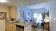 This single-floor residence offers a thoughtfully designed open-concept layout that seamlessly integrates the kitchen and living areas into one cohesive, functional space. The kitchen features light wood cabinetry, a granite-topped breakfast bar, a stainless steel sink, and a built-in dishwasher, making it both practical and visually appealing. The kitchen flows effortlessly into the living area, which is distinguished by a calming light blue accent wall adorned with white wall-mounted shelving units that provide stylish display and storage space. The living area is comfortably appointed and benefits from an abundance of natural light streaming in through a large window dressed with sheer, patterned curtains. Parquet-style flooring adds warmth and character to the living space, while the vaulted ceiling height enhances the sense of openness and airiness throughout. A floor lamp and a neatly arranged desk area contribute to the room's versatility, supporting both relaxation and productivity. Lush greenery near the window adds a refreshing touch of nature to the interior. Overall, the space presents a welcoming, bright, and well-organized environment with a clean, contemporary aesthetic.