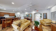 Located at Hawks Landing Drive in Fort Myers, Florida, this single-floor community clubhouse offers a thoughtfully designed layout across a generous footprint. The entrance welcomes visitors with soaring double-height ceilings, floor-to-ceiling windows that flood the space with natural light, and warm earth-toned tile flooring laid in a classic diagonal pattern throughout the main areas. An elegant arched passageway connects the main living room to additional spaces, creating a seamless flow across the facility. The primary living room is bright and open, furnished with comfortable seating and accented by large multi-pane windows that frame views of the surrounding Florida landscape. A combined kitchen and living area features a substantial wrap-around wooden bar counter, rich cabinetry, and full appliances, ideal for gatherings and events. Two dedicated office spaces serve administrative needs—one outfitted as a multi-workstation computer lab with built-in desks and printers, and another configured as a private management office. The game room provides a lively retreat, complete with a full-size pool table, vintage-style artwork, and light wood flooring. Ceiling fans, recessed lighting, and lush indoor greenery throughout contribute to the relaxed, welcoming ambiance of this well-maintained Florida clubhouse.