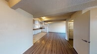 This single-floor space offers a practical and open layout, with the kitchen and living area flowing seamlessly into one another. The kitchen is well-equipped with white cabinetry, dark countertops, a double-basin sink, and white appliances including a range, dishwasher, and refrigerator. Light tile flooring defines the kitchen zone, while warm hardwood-style flooring extends throughout the main living area, creating a clear visual transition between spaces.

The living area is generously proportioned, featuring a textured ceiling with exposed structural beams that add architectural character. Flush-mount light fixtures provide overhead illumination throughout, and baseboard heaters run along the walls. A window allows natural light into the kitchen, and a door provides direct access to an outdoor area with views of surrounding trees.

A walk-in storage or closet area is accessible from the living space, offering additional utility. The bathroom includes a full tub and shower combo with white tile surround, a toilet, and a frosted window that allows natural light while maintaining privacy. The overall palette is neutral throughout, with white walls and trim contributing to a clean, functional aesthetic.