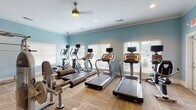 This fitness center offers a comprehensive workout environment housed within a single-floor facility. The space is finished with soft blue walls, white crown molding, and a parquet-style wood-look flooring that lends a clean, welcoming aesthetic throughout.

The cardio area is well-appointed with multiple treadmills and elliptical machines, each equipped with individual monitor screens for entertainment during workouts. A ceiling fan overhead aids in air circulation, complementing the recessed lighting that keeps the space bright and energizing. Large windows fitted with white blinds line the walls, allowing ample natural light to flow in while providing views to the outdoors.

The strength training section features a multi-station cable machine, a squat rack, a weight bench, and a dedicated free weights area with a full rack of dumbbells. A tiered medicine ball rack holds a varied selection of weighted balls, catering to functional fitness and cross-training routines.

Full-length mirrors are strategically positioned along one wall, expanding the visual sense of the space and allowing users to monitor their form during exercise. A stationary recumbent bike and additional resistance machines round out the equipment offerings. Access doors with glass panels connect the space to adjacent areas, contributing to an open, airy feel throughout this thoughtfully outfitted fitness facility.