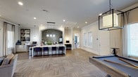 This single-floor communal space is designed with a clean, contemporary aesthetic that balances social and functional areas seamlessly. The open-plan layout allows for a natural flow between zones, with wood-look parquet-patterned flooring grounding the interior in warm, earthy tones throughout.

A central bar-style counter serves as the focal point of the space, framed by an elegant arched alcove clad in a dark mosaic tile backsplash that adds texture and visual depth. Pendant lighting hangs above the counter, complementing the recessed ceiling lights that illuminate the room evenly. Navy blue upholstered bar stools line the counter, offering casual seating for socializing or dining.

Adjacent to the bar area, a built-in workstation with a monitor provides a functional workspace nook, ideal for residents needing a quiet area for focused tasks. On the opposite side of the room, a pool table introduces a recreational element, reinforcing the space's role as a social hub.

White crown molding, crisp white walls, and paneled doors contribute to the polished, upscale finish of the interior. The overall ambiance is welcoming and versatile, suited for both relaxed gatherings and productive use.