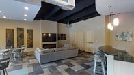 This single-floor space showcases a striking industrial-modern aesthetic defined by its soaring ceilings with exposed black-painted ductwork and ventilation systems, creating a bold architectural contrast against the neutral, light-toned walls. The main lounge area is anchored by a built-in linear electric fireplace set beneath a wall-mounted flat-screen television, flanked by open shelving niches that add both function and visual interest. A large, sectional sofa in a cool grey tone provides ample seating and sits atop a geometric, multi-toned carpet that adds texture and depth to the floor. To the right, bar-height seating with metallic stools hints at a transitional dining or social area, further emphasizing the space's communal character. Warm-toned wooden doors provide a natural contrast to the otherwise industrial palette. The generous ceiling height amplifies the sense of openness throughout, while the mix of raw industrial elements and polished furnishings creates a cohesive, contemporary environment well-suited for social gatherings or relaxed lounging.