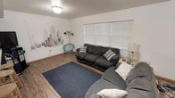 This single-floor space centers around a comfortable living area that combines casual, functional design with subtle decorative touches. The room features warm wood-look laminate flooring throughout, providing a cohesive and easy-to-maintain surface underfoot. White walls serve as a clean backdrop, with a standout hand-painted or printed cactus and desert botanical mural adorning one wall, adding a distinctive southwestern artistic element to the space.

Natural light enters through a large window fitted with horizontal white blinds, offering both brightness and privacy control. Overhead, a flush-mount ceiling light provides additional illumination for evening use. The living area comfortably accommodates a full-sized dark gray sofa and a matching sectional or additional sofa piece, arranged around a navy blue area rug that anchors the seating arrangement. A small turquoise accent chair adds a pop of color alongside the mural. A side table with a lamp completes the setup, adding to the room's relaxed and lived-in feel. The overall ambiance is casual and welcoming, with eclectic décor choices that give the space a personalized character.