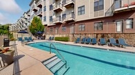 Located at 970 Riverside Forest Way in Knoxville, Tennessee, this thoughtfully designed outdoor amenity space offers a comprehensive resort-style experience set against a scenic riverfront backdrop. The centerpiece is a generously sized swimming pool with crystal-clear blue water, surrounded by a spacious pool deck furnished with chaise lounges for relaxed sunbathing. Tiered stone retaining walls add architectural depth, while the multi-story residential building rises impressively overhead, with private balconies overlooking the amenity area below.

A well-appointed outdoor kitchen features built-in grills and prep stations, shaded by large navy umbrellas, making it ideal for entertaining. Nearby, a cozy fire pit seating area with Adirondack chairs in bold blue provides an inviting gathering spot. Wide concrete pathways connect each zone seamlessly, flanked by vibrant landscaping that includes ornamental grasses, cypress trees, flowering plants, and decorative teal ceramic planters that add pops of color throughout.

The overall ambiance is lively yet relaxed, blending functional amenities with natural surroundings. Mature trees and greenery frame the space, offering shade and privacy while maintaining open views toward the river. The combination of brick and lap-siding exterior architecture gives the property a refined, contemporary character that complements its natural riverside setting.