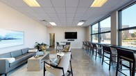 Located at 814 N 3rd St in Wilmington, NC, this single-floor space offers a thoughtfully designed and versatile layout suited for gathering, socializing, or focused work. The open main area is anchored by a comfortable lounge zone furnished with a contemporary gray sofa, accent chairs, and a concrete coffee table set atop a soft area rug, creating an inviting, relaxed atmosphere. A wall-mounted flat-screen television with a soundbar serves as a central media focal point.

One of the most striking architectural features is the expansive floor-to-ceiling window wall that lines one side of the space, flooding the interior with abundant natural light while offering clear views of the surrounding Wilmington streetscape. Running along this window wall is a long high-top counter with bar-height seating, ideal for individual work or informal collaboration.

Tile flooring extends throughout the main area, providing a clean, durable foundation that complements the neutral, modern color palette. Recessed panel ceiling tiles with integrated lighting ensure consistent, even illumination throughout.

A dedicated kitchenette is equipped with rich dark cabinetry, granite countertops, a mosaic tile backsplash, a refrigerator, and ample storage — making it a fully functional support space. Together, these elements combine to create a polished, multi-use environment well-suited for community or professional use.