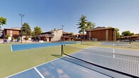 This outdoor recreational facility centers around a well-appointed complex of tennis courts, offering ample space for players of all skill levels. The courts feature a striking dual-tone surface design, with blue and green hard-court finishes clearly marked with crisp white boundary lines. Each court is equipped with regulation nets and is surrounded by sturdy black chain-link fencing to define play areas and ensure safety.

The aerial view reveals multiple full-sized courts arranged side by side, maximizing the available space and accommodating simultaneous matches. Tall light poles are positioned throughout the complex, suggesting the facility supports evening play. A separate, smaller court with a solid green surface is also part of the complex, offering additional variety.

Adjacent to the courts, a swimming pool area with lounge chairs and sun umbrellas provides a complementary recreational amenity. A two-story clubhouse building with an upper-level deck overlooks the courts and pool, offering a convenient gathering space. The surrounding landscape features mature trees and well-maintained green lawns, lending a park-like ambiance to the facility. Brick residential buildings frame the perimeter, situating the courts within a larger community setting under open, sunny skies.