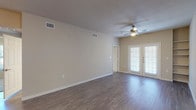 This single-story residence offers a well-organized layout with distinct living, sleeping, and functional zones. The main living area is a generously proportioned room finished with dark hardwood-style flooring and neutral beige walls. A ceiling fan with a light fixture is centrally positioned overhead, and a pair of French doors along one wall allows natural light to filter in while providing access to the exterior. A HVAC vent and multiple electrical outlets confirm the room's practical infrastructure.

The kitchen is efficiently arranged in an L-shape configuration, featuring light maple cabinetry both above and below the countertops, a speckled laminate countertop surface, a double stainless steel sink, a dishwasher, an electric range with overhead microwave, and a full-size refrigerator. A pass-through view connects the kitchen directly to the living area.

The bedrooms are carpeted and include ceiling fans and built-in wall shelving with a small desk surface, offering functional storage solutions. Natural light enters through windows fitted with blinds.

Two bathrooms are present, each featuring a tub-shower combination with white tile and gray accent tiles, wood-style flooring, and light maple vanity cabinetry. One bathroom includes accessibility grab bars near the tub.