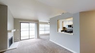 This single-floor space offers an open and welcoming layout designed to maximize both comfort and functionality. The living area is finished with plush wall-to-wall carpeting in a neutral tone, creating a warm and inviting atmosphere. Natural light floods the space through a large sliding glass door and an adjacent wide window, offering outdoor views and a strong connection to the exterior environment. A ceiling fan is centrally positioned above the living area, adding a practical element to the room's clean design.

A defining architectural feature is the pass-through window cut into the dividing wall between the living space and the kitchen. This thoughtful design element opens the two areas to one another, promoting an airy, connected feel without fully removing the separation between spaces. The kitchen, visible through the pass-through, is outfitted with white cabinetry, a built-in microwave, and a range, providing a functional cooking environment. The countertop extends to form a convenient ledge accessible from the living side.

The overall color palette is light and neutral throughout, with soft gray walls and white trim contributing to a clean, contemporary ambiance. The space feels well-proportioned and efficiently laid out.