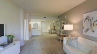 This single-floor space offers an open-concept layout that connects the living, dining, and kitchen areas in a fluid, functional arrangement. The living area features neutral-toned carpet flooring and warm, light-colored walls that create a bright and welcoming ambiance. A comfortable seating area anchors the space, complemented by a white open-frame shelving unit that adds both storage and visual interest. The dining area sits adjacent to the living space, furnished with a modest table and bench seating, with a decorative mirror adding depth to the room. Track lighting overhead ensures the area is well illuminated. The kitchen is accessible directly from the main living area and features white cabinetry, a raised breakfast bar with white barstools, and pendant lighting that adds a modern touch. The overall color palette is cohesive, relying on soft grays, whites, and warm neutrals to create a calming, contemporary feel. Recessed ceiling vents are visible throughout, indicating a central climate control system. The open layout maximizes the sense of space, allowing natural sightlines across the entire floor plan, making the interior feel airy and well-proportioned.