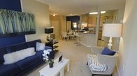 This single-floor residence offers a thoughtfully designed open-concept layout that integrates the living, dining, and kitchen areas into one cohesive and functional space. The living area is anchored by a bold navy blue sofa accented with decorative pillows, complemented by a sleek white coffee table and contemporary accent chairs. An accent wall in deep navy adds a striking visual focal point, lending the space a modern and polished character.

The dining area sits just beyond the living space, featuring a stylish table with upholstered seating, ideal for both casual meals and entertaining. A breakfast bar with bar stools creates a natural transition into the kitchen, which is equipped with warm wood cabinetry, a white refrigerator, and ample counter space. The kitchen's efficient layout ensures ease of movement and accessibility.

Throughout the space, neutral-toned flooring ties the rooms together, amplifying the sense of openness and continuity. Recessed and overhead lighting provides consistent illumination across the entire floor plan. The overall ambiance is clean, contemporary, and welcoming, with a careful balance of bold accents and neutral tones that create a stylish yet comfortable living environment.