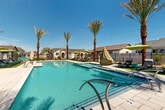 Located in Phoenix, Arizona, this outdoor community amenity area offers a resort-style experience designed for relaxation and recreation. The centerpiece is a generously sized swimming pool with crystal-clear blue water, complemented by a separate spa with tiled surrounds and built-in entry rails. The pool deck is finished in light-toned pavers, providing ample space for sunbathing and socializing. Tall, mature palm trees are strategically placed throughout the area, adding a tropical ambiance and natural shade under the abundant Arizona sun. A dedicated lounge zone features multiple chaise loungers and seating arrangements shaded by vibrant green market umbrellas, creating comfortable spots to unwind poolside. The entire area is enclosed by a wrought-iron style perimeter fence, ensuring privacy and security. A utility and storage structure is situated at one end of the deck, keeping the space organized and functional. Patches of artificial turf add a touch of greenery between seating areas. The surrounding neighborhood consists of single-story residential homes with pitched rooflines, visible just beyond the fencing. Under the expansive blue Phoenix sky, this well-appointed outdoor space delivers a welcoming, resort-inspired atmosphere for its community residents.