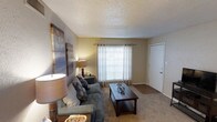 This single-floor residential space offers a warm and functional layout designed for comfortable everyday living. The living room serves as the heart of the home, featuring wall-to-wall carpeting that adds a soft, cozy feel underfoot. A large window dressed with horizontal blinds and sheer curtains allows an abundance of natural light to fill the room, creating a bright and welcoming atmosphere. The textured popcorn ceiling adds a classic character to the space. Neutral-toned walls provide a versatile backdrop, complementing a range of décor styles. The room offers ample space for seating and a central coffee table, making it well-suited for both relaxing and entertaining. A doorway visible within the living area hints at additional rooms beyond, suggesting a thoughtful and connected floor plan throughout the residence. Overall, the space presents a practical and inviting environment with a straightforward, residential aesthetic that balances functionality with comfort.