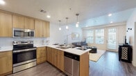 This single-floor residence is designed with a contemporary aesthetic, combining functionality and style across its well-appointed layout. The open-concept kitchen and living area form the heart of the space, where light wood cabinetry, white quartz countertops, and a full suite of stainless steel appliances — including a refrigerator, range, microwave, and dishwasher — create a clean, modern kitchen. A central island with pendant lighting overhead serves as both a prep area and casual dining space with seating. Wood-look flooring runs throughout the main living areas, adding warmth and continuity.

The living room benefits from an abundance of natural light, courtesy of large windows and a glass door that opens to an outdoor patio area. Neutral walls and recessed lighting contribute to the bright, airy atmosphere of the space.

The bedroom is carpeted for comfort and includes a ceiling fan, windows with views to the exterior, and direct access to a spacious walk-in closet with built-in shelving. The full bathroom features a wood-toned vanity with a wide mirror, white quartz countertop, and a combined tub and shower with wood-look flooring completing the room's clean, cohesive design.