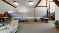 Located at 440 River Rd in Manchester, NH, this spacious single-floor loft offers a generously sized, open-concept layout defined by striking architectural character. Prominent dark wood beams run throughout the vaulted, angled ceilings, lending a warm rustic charm that contrasts beautifully with the neutral-toned walls and plush wall-to-wall carpeting underfoot.

The loft is thoughtfully arranged into distinct functional zones. A dedicated game area anchors one section of the space, featuring a full-size pool table beneath a classic dome pendant light and a ping-pong table illuminated by a multi-shade hanging fixture — ideal for recreation and entertainment. Exposed brick accent walls add texture and warmth to this area, complemented by a window that draws in natural light.

Adjacent to the game zone, a comfortable lounge area provides seating with soft furnishings and a wall-mounted television, creating an inviting space for relaxation. A dining area with a large table and upholstered chairs sits beneath skylights that flood the room with daylight, enhancing the airy, open ambiance.

Multiple skylights and flush ceiling fixtures ensure consistent, well-distributed lighting throughout. The overall atmosphere of this loft is both functional and characterful — a versatile, amenity-rich space well-suited for gathering, leisure, and everyday use.
