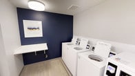 This shared laundry room combines functionality with a polished aesthetic. The space is defined by a striking navy blue accent wall, which adds a bold contrast to the surrounding white walls and creates a visually engaging focal point. A framed piece of abstract artwork hangs on the accent wall, contributing a decorative touch that elevates the room beyond purely utilitarian design. The flooring consists of light wood-look planks, lending warmth and a contemporary feel to the space. Multiple top-loading washing machines with digital payment interfaces are neatly arranged along one wall, offering convenient, modern coin-free or card-based operation. A wall-mounted folding shelf provides a practical surface for sorting or folding laundry. Overhead, a flush-mount ceiling light provides bright, even illumination throughout the room. The textured ceiling and ventilation grille indicate thoughtful infrastructure planning. Overall, the laundry room is well-maintained, efficiently organized, and designed with both practicality and visual appeal in mind, making it a comfortable and functional amenity.