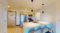 This contemporary single-floor residence is designed with a clean, modern aesthetic and an efficient open layout. The kitchen is a standout feature, showcasing light wood cabinetry, stainless steel appliances including a refrigerator, microwave, and range, complemented by sleek white quartz countertops. A large kitchen island with an undermount sink and bar seating serves as both a functional workspace and a casual dining area, illuminated by stylish dark pendant lights overhead. Light wood-toned flooring flows throughout the space, enhancing the bright and airy atmosphere created by recessed lighting.

The living area offers a comfortable, minimalist setting with a leather sofa and exposed concrete ceiling accents, lending an industrial touch to the modern design. Windows in the living and bedroom areas frame views of the surrounding urban landscape, filling the rooms with natural light.

The bedroom provides a quiet retreat with a ceiling fan, ample natural light, and space for a dedicated work area. The bathrooms are finished with white quartz countertops, light wood vanity cabinetry, and large mirrors. One bathroom features a dual-sink vanity, offering added convenience. A separate water closet adds privacy and functionality. The overall design is cohesive, combining contemporary finishes with practical layouts to create a well-appointed living environment.