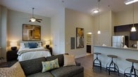 This single-floor studio is designed with an open-concept layout that efficiently integrates the living, sleeping, and kitchen areas into one cohesive space. Notably high ceilings lend a sense of spaciousness and airiness throughout. The living area features a charcoal upholstered sofa accented with decorative throw pillows, positioned beneath a ceiling fan that complements the vertical height of the room. Hardwood-style flooring runs continuously throughout, adding warmth and visual continuity.

The sleeping area is defined by a ceiling fan, a well-appointed bed with layered bedding, and a bedside lamp providing ambient lighting. Abstract artwork adorns the walls, adding color and character to the neutral palette of soft greens and whites.

Adjacent to the main living space, the kitchen area includes a peninsula counter with bar seating, pendant lighting overhead, and light-toned countertops. A glimpse into the bathroom reveals decorative tile work and cabinetry. The overall ambiance is modern yet inviting, with warm lighting and thoughtful design elements throughout.