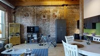 Located at 300 Blackwell St in Durham, NC, this single-floor loft showcases the character and craftsmanship of a thoughtfully preserved industrial space. The open-plan layout is anchored by dramatic exposed brick walls that span the full height of the interior, bearing the patina of age and history — including ornate arched brick detailing that adds a distinctly architectural touch. Heavy timber ceiling beams run overhead, complementing the warm, light-toned hardwood floors that extend throughout the space, creating a cohesive and inviting aesthetic.

The living area flows naturally into a dining zone, with the open layout allowing for flexible use of the generous floor space. Structural columns punctuate the interior, serving as a reminder of the building's industrial heritage while adding visual rhythm to the layout. Exposed conduit and raw building materials are seamlessly integrated into the design, lending an authentic loft character throughout.

Natural light filters into the space, highlighting the rich texture of the brick and the warmth of the wood elements. Storage is thoughtfully incorporated with cabinetry that complements the moody, earthy tones of the interior. Overall, the space strikes a balance between rugged industrial charm and comfortable, livable warmth.