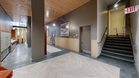 Located in Portland's vibrant Pearl District, this ground-floor lobby at 123 NW 12th Ave offers a generously scaled, design-forward communal space. The entrance is anchored by floor-to-ceiling glass doors and curtain walls that flood the interior with natural light and provide open views of the surrounding streetscape. Soaring double-height ceilings with warm wood slat paneling create a striking architectural contrast against the polished concrete and herringbone-patterned light wood flooring that define the circulation path throughout.

The space is organized into several distinct zones. A welcoming lounge near the entrance features richly upholstered burnt-orange seating and layered textiles, while a pair of retro-inspired armchairs in a quieter alcove offer a more intimate setting. A large communal dining table with sculptural wooden legs serves as a central gathering point, surrounded by artisan chairs beneath pendant lighting. A glass-walled private meeting room provides a semi-transparent enclosure, maintaining visual connectivity while offering separation. A staffed reception desk with warm wood paneling sits adjacent to the staircase. The overall palette of muted gray, terracotta, and natural wood tones, complemented by curated artwork throughout, gives the lobby a cohesive, sophisticated, and welcoming ambiance.