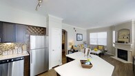 Located at 8501 East Alameda Avenue in Denver, Colorado, this single-floor residence offers a thoughtfully designed open-concept layout that maximizes both functionality and comfort. The kitchen serves as a central hub of the home, outfitted with stainless steel appliances, including a full-size refrigerator, and a spacious white countertop island that provides ample prep space and casual seating. Dark cabinetry provides a striking contrast to the bright white surfaces, while a decorative wine rack adds a charming, custom touch. Crown molding and clean white walls lend the space a polished, refined character throughout. An elegant arched doorway connects the kitchen to the living area, creating a natural flow between spaces. The living room is warm and inviting, furnished with a comfortable sofa accented by cheerful yellow and blue throw pillows, and is anchored by a soft lamp that adds ambient lighting. A dining area sits adjacent, bathed in natural light from a large window fitted with blinds. Hardwood-style flooring runs consistently throughout, unifying the open floor plan. The overall ambiance is bright, modern, and welcoming, striking a balance between stylish design and everyday livability.