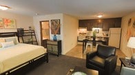 This single-floor studio-style space offers an efficient and thoughtfully arranged open-plan layout that seamlessly integrates the sleeping, living, and kitchen areas. The living and sleeping zone features carpeted flooring, providing a warm and comfortable feel, and is furnished with a full-sized bed and a leather armchair, making it both functional and inviting. A decorative mosaic-front dresser and wall art add personality and a touch of eclectic style to the space. The kitchen area transitions smoothly from the living zone via a tile floor and features dark espresso-toned cabinetry, a breakfast bar with metal stools, a microwave, and under-cabinet lighting that creates a warm ambiance. Full appliances are included, making the kitchen fully operational. A partial wall divides the sleeping area from the kitchen, offering a sense of separation without closing off the open feel of the space. A doorway leads to a private bathroom, visible in the background, which adds to the self-contained nature of the unit. The textured ceiling and neutral wall tones throughout contribute to a clean, cohesive aesthetic. Overall, the space is compact yet efficiently designed to accommodate everyday living comfortably.