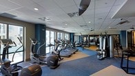 This fitness center is designed to accommodate a wide range of workout styles and training needs within a single, open-plan floor. The space is appointed with a drop-panel ceiling fitted with recessed lighting and ceiling fans, ensuring consistent illumination and airflow throughout workouts. Large windows along the perimeter walls allow natural light to fill the room, creating a bright and energizing environment.

The cardio zone features a row of treadmills, elliptical trainers, and upright exercise bikes, all positioned to take advantage of the natural light. A wall of mirrors extends along one side of the space, visually expanding the room and allowing users to monitor their form. The strength training area is equipped with a multi-station cable and weight rack system, along with a dedicated free weights section that includes a full set of dumbbells, kettlebells, and medicine balls organized on tiered racks.

The functional training zone provides stability balls, resistance bands, and TRX suspension trainers, offering versatile options for dynamic and bodyweight-focused exercises. The flooring transitions between cushioned carpet tiles and a hard, smooth surface, delineating workout zones while providing appropriate support. Bold blue accent walls add a modern, motivating aesthetic to the overall design of the space.