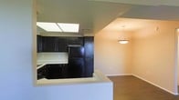 This single-story space showcases an open-concept layout where the kitchen seamlessly connects to the dining area, creating a cohesive and functional living environment. The kitchen is outfitted with dark espresso-toned cabinetry that provides a bold contrast against the light countertops, lending the space a classic yet striking aesthetic. Black appliances, including a refrigerator and range, complement the cabinetry for a cohesive look. A notable architectural feature is the skylight positioned above the kitchen, which floods the cooking area with natural light and enhances the sense of openness. The kitchen's peninsula-style counter offers additional workspace and serves as a natural divider between the kitchen and the adjoining dining area. Transitioning into the dining space, warm hardwood-style flooring adds texture and warmth underfoot. A pendant light fixture hangs centrally over the dining area, providing focused ambient lighting. The neutral-toned walls throughout contribute to a clean, inviting atmosphere, while the open sightlines between the kitchen and dining area make the overall space feel expansive and well-connected.