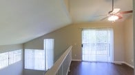 This two-story space showcases a well-designed upper level loft area that highlights several architectural details. The upper floor features striking vaulted ceilings that angle upward, creating an open and airy atmosphere. Dark hardwood flooring adds warmth and contrast against the light-toned walls throughout the space.

A white-railed open staircase connects the two floors, contributing to the sense of openness by allowing sightlines between levels. Vertical blinds dress both the stairwell-adjacent window and a full sliding glass door, which serves as a prominent feature of the upper level. The sliding door floods the space with abundant natural light while offering views of the greenery outside, seamlessly connecting the interior to the outdoors.

The ceiling features a recessed panel detail and a ceiling fan, adding both functional and visual interest. Neutral wall tones complement the overall design, lending the space a clean, versatile aesthetic. Together, the vaulted ceilings, natural light, and open layout create a bright, inviting environment that feels spacious and well-connected across both levels.