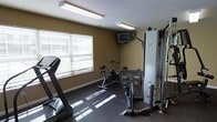 This single-floor fitness room is designed to provide a functional and accessible workout environment. The space features warm-toned walls paired with dark rubberized flooring, creating a practical and focused atmosphere suited for exercise. Natural light filters in through a large window dressed with horizontal blinds, brightening the room and offering a pleasant contrast to the overhead fluorescent lighting.

The room is outfitted with a selection of cardio and strength-training equipment, including a treadmill, a stationary spin bike, and an elliptical-style machine, catering to a variety of fitness routines. A multi-station cable weight machine positioned along one wall provides options for resistance and strength training. A wall-mounted television is conveniently placed within view of the equipment, allowing users to stay entertained during their workouts.

The layout makes efficient use of the available space, arranging the equipment to allow for comfortable movement between stations. White painted baseboards and clean walls contribute to a tidy, well-maintained appearance. Overall, this fitness room serves as a straightforward and fully equipped space for individuals looking to maintain an active lifestyle.