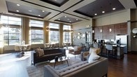 This elegantly designed single-floor space showcases a seamless blend of contemporary style and functional comfort. The centerpiece of the main living area is a striking coffered ceiling with dark-framed geometric panels and recessed lighting, lending a sense of architectural depth and grandeur. Large floor-to-ceiling windows line the walls, flooding the interior with natural light while offering views of the surrounding exterior landscape. Sheer gold-toned drapery frames the windows, adding warmth and a refined aesthetic.

The seating area is thoughtfully arranged with a tufted light-gray sectional, a dark leather sofa, and transitional accent chairs, all anchored by a patterned area rug. Decorative pendant chandeliers with a globe-like design contribute an artistic focal point overhead. A sunburst mirror and circular wall art adorn one wall, enhancing the space's visual interest.

Toward the rear, a bar or café-style counter with bar seating and dark cabinetry suggests a hospitality or amenity function, complemented by pendant lighting and appliances. The overall ambiance is upscale, welcoming, and ideal for both social gatherings and relaxed lounge settings.