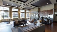 This elegantly designed single-floor space showcases a seamless blend of contemporary style and functional comfort. The centerpiece of the main living area is a striking coffered ceiling with dark-framed geometric panels and recessed lighting, lending a sense of architectural depth and grandeur. Large floor-to-ceiling windows line the walls, flooding the interior with natural light while offering views of the surrounding exterior landscape. Sheer gold-toned drapery frames the windows, adding warmth and a refined aesthetic.

The seating area is thoughtfully arranged with a tufted light-gray sectional, a dark leather sofa, and transitional accent chairs, all anchored by a patterned area rug. Decorative pendant chandeliers with a globe-like design contribute an artistic focal point overhead. A sunburst mirror and circular wall art adorn one wall, enhancing the space's visual interest.

Toward the rear, a bar or café-style counter with bar seating and dark cabinetry suggests a hospitality or amenity function, complemented by pendant lighting and appliances. The overall ambiance is upscale, welcoming, and ideal for both social gatherings and relaxed lounge settings.