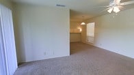 This single-floor residential space offers a straightforward, functional layout designed for comfortable everyday living. The interior is finished with neutral-toned carpeting throughout, creating a cohesive and understated aesthetic across the living and dining areas. The open floor plan allows for a seamless flow between spaces, with a partial dividing wall subtly defining zones without fully enclosing them. A ceiling fan is positioned overhead in the living area, providing year-round climate comfort. The dining area is anchored by a classic pendant light fixture that hangs from the ceiling, adding a warm, focused glow to the space. Natural light filters in through a window fitted with horizontal blinds, brightening the dining zone and contributing to an airy atmosphere. Adjacent to the dining area, a pass-through or countertop area hints at a connected kitchen space, offering convenience for daily routines. The walls are painted in a clean, light neutral tone, enhancing the sense of openness. HVAC vents are integrated into the ceiling and walls, ensuring consistent airflow. Overall, the space presents a clean, move-in ready environment with a calm and practical ambiance.
