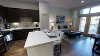 This well-appointed single-floor residence showcases a thoughtfully designed open-concept layout that seamlessly connects the kitchen, dining, and living areas into one cohesive and functional space. The kitchen is outfitted with rich dark cabinetry, a neutral tile backsplash, stainless steel appliances, and a deep undermount sink, offering both style and practicality. A large white kitchen island anchors the space, providing additional seating with modern high-back chairs — ideal for casual dining or entertaining. The dining area flows naturally from the kitchen, maintaining the open and airy feel throughout. The living room is warmly furnished and benefits from natural light filtering through window blinds, creating a comfortable and inviting ambiance. Rich, dark hardwood-style flooring runs throughout the entire space, adding warmth and visual continuity. Neutral gray walls serve as a refined backdrop, complemented by carefully selected décor including accent lighting, teal throw pillows, and a patterned area rug. A tufted leather ottoman adds a touch of sophistication to the living area. Overall, the space balances modern aesthetics with functional design, making it a comfortable and stylish environment.