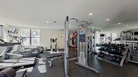 This single-level athletic center offers a thoughtfully organized fitness facility designed to accommodate a wide variety of workout routines. The interior features white walls, recessed lighting, and dark rubber flooring throughout, creating a clean, functional environment conducive to exercise.

The strength training area is well-appointed with a cable crossover machine, free weight benches, and a full dumbbell rack ranging across multiple weight increments. A dedicated functional training zone includes a heavy punching bag, stability balls, and medicine balls arranged on a storage rack, providing options for dynamic and cross-training workouts.

The cardio section is positioned near large windows that invite natural light and outdoor views, housing treadmills, an elliptical trainer, and stationary cycling bikes. A wall-mounted television provides entertainment for cardio users.

Floor-to-ceiling mirrors line key sections of the walls, visually expanding the space and allowing users to monitor their form. The facility is accessed through glass doors that further enhance the sense of openness. Artwork is displayed near the entrance, adding a polished touch to the athletic environment. Posted facility policies indicate structured management of the space.