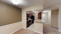 This single-story residence at Oldham Oaks offers a practical and cohesive layout designed for comfortable everyday living. The living area is finished with wall-to-wall carpeting in a neutral tone, complemented by warm beige walls and crisp white chair-rail molding that adds a subtle architectural detail along the lower portion of the walls. Flush-mount ceiling lighting provides consistent illumination throughout. The kitchen is accessible through an open arched passageway, creating a seamless connection between the living and cooking spaces. It features rich dark wood cabinetry that contrasts with light-toned countertops, and is equipped with black appliances including a refrigerator, range, and over-the-range microwave. The kitchen floor is finished with light-toned tile, distinguishing it from the carpeted living area. A peninsula with a granite-style countertop extends into the transitional space, offering additional prep or dining space. A doorway visible beyond the kitchen suggests further rooms within the unit. The textured ceiling throughout adds depth to the overall interior finish. The space presents a clean, move-in-ready environment with a warm and functional character.