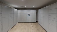 The Package Concierge room is a single-floor, purpose-built space designed to streamline parcel management for residents. The room is defined by an extensive array of floor-to-ceiling automated lockers finished in a uniform light gray, lining multiple walls and offering a wide range of compartment sizes to accommodate packages of varying dimensions — from small envelopes to large deliveries. A centrally mounted digital touchscreen interface on the locker bank serves as the primary point of interaction, allowing residents to receive notifications and retrieve their packages securely and efficiently. The flooring consists of polished terrazzo-style concrete, lending the room a clean, utilitarian aesthetic. Recessed ceiling lights provide consistent, even illumination throughout the space. A glass-paneled door, clearly labeled "Package Concierge," provides access to the room and offers a view into an adjacent common area beyond. A security camera is also present, enhancing the overall safety of the space. The room's straightforward, well-organized layout prioritizes functionality, ensuring a seamless package retrieval experience.